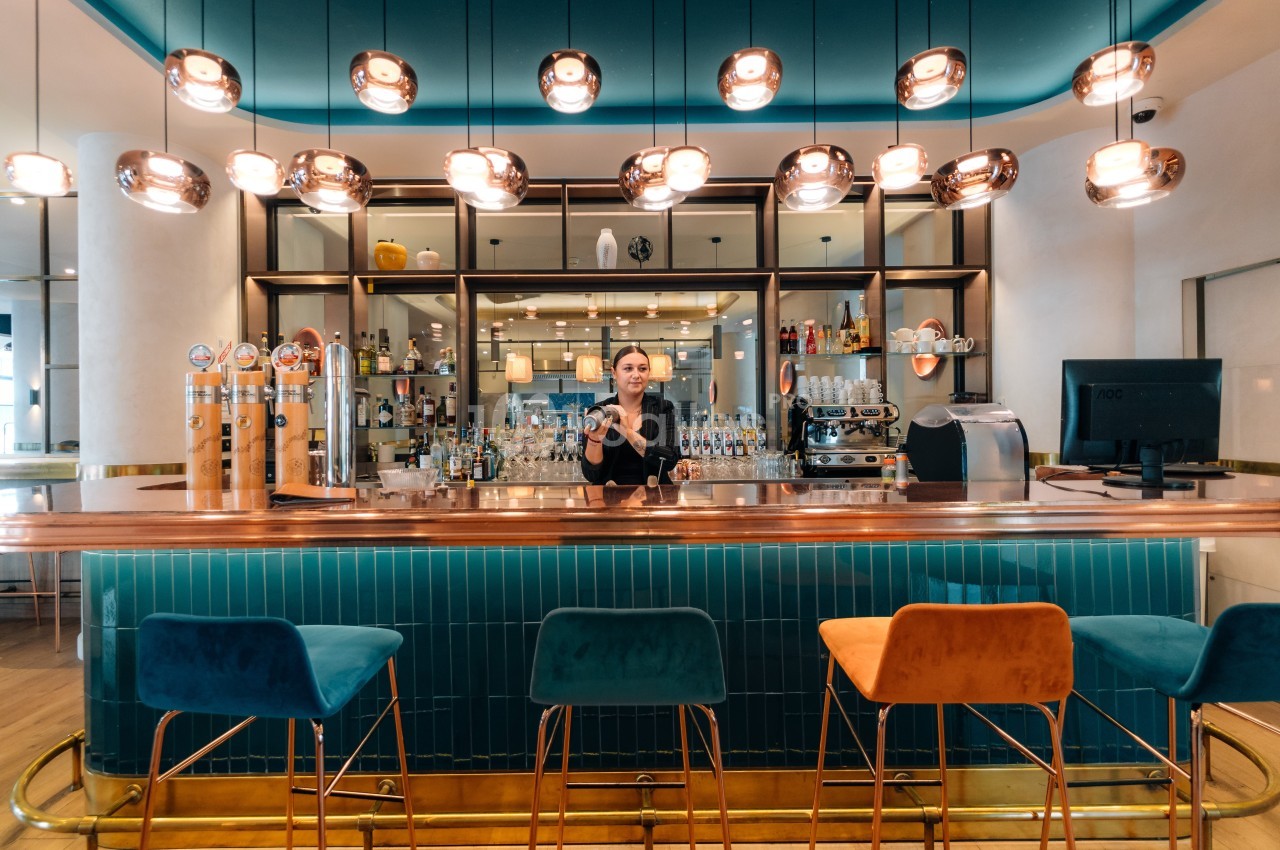Un barman derrière un comptoir en bois dans un bar moderne avec des tabourets colorés et des luminaires suspendus.