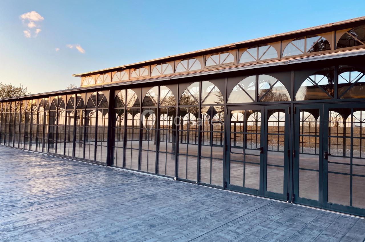 Grande serre en verre avec structure métallique et portes vitrées, reflétant le ciel au coucher du soleil.