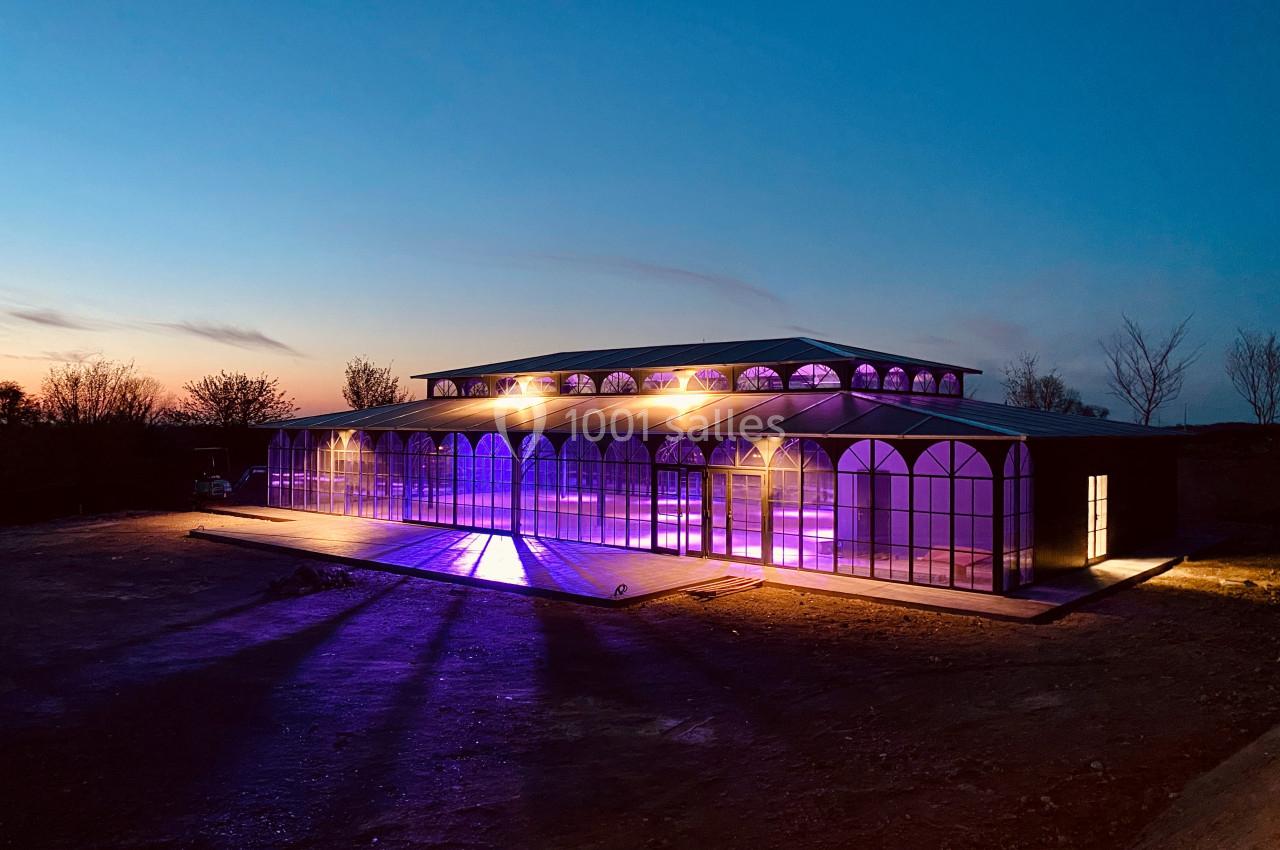 Bâtiment vitré illuminé par des lumières violettes et jaunes au crépuscule, entouré d'un terrain dégagé.