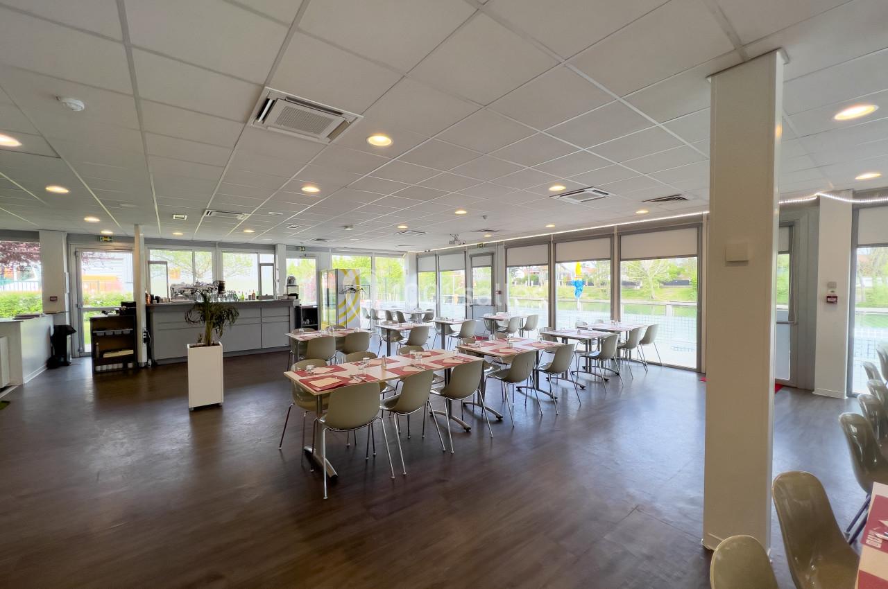 Salle de restaurant lumineuse avec grandes baies vitrées, tables dressées et chaises modernes sur un sol en bois sombre.