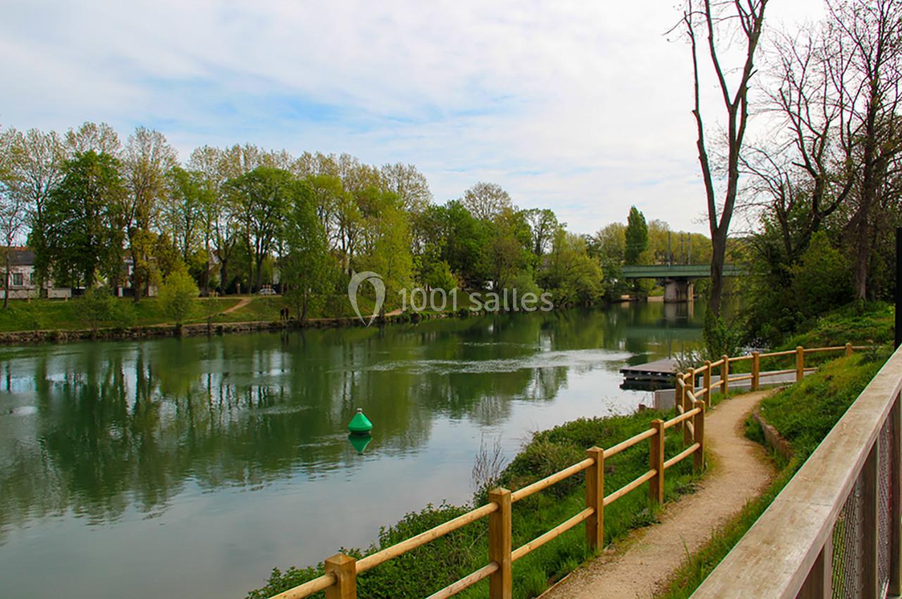 Chemin bordé de barrières en bois longeant une rivière calme avec des arbres et un pont en arrière-plan.