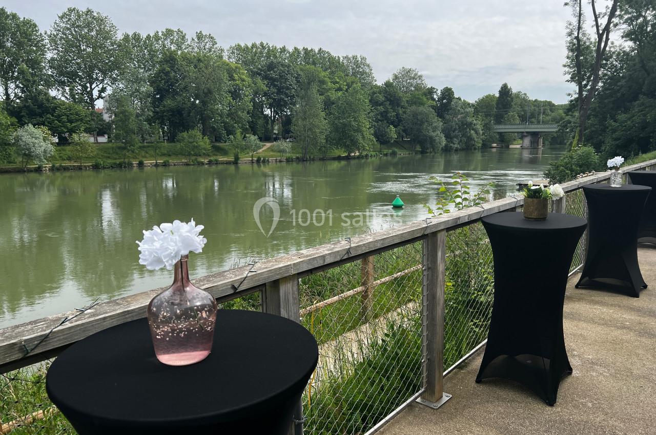 Tables hautes avec nappes noires et vases fleuris sur une terrasse surplombant une rivière bordée d'arbres.