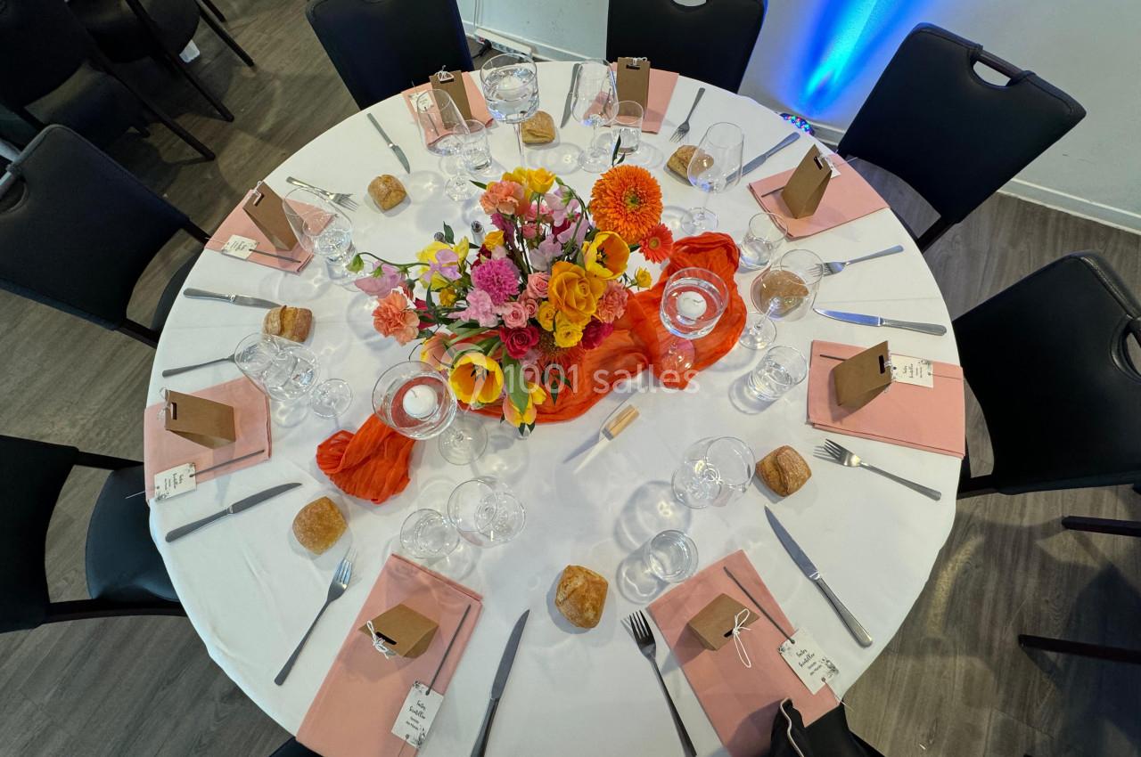Table ronde dressée avec une nappe blanche, centre de table floral coloré, serviettes roses et couverts disposés.