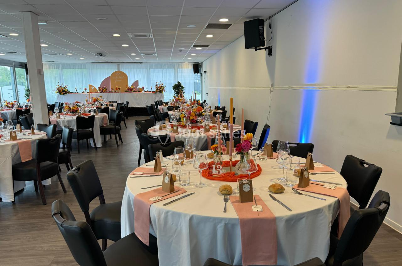 Salle de réception décorée avec des tables rondes dressées, nappes blanches, chemins de table roses et arrangements floraux.