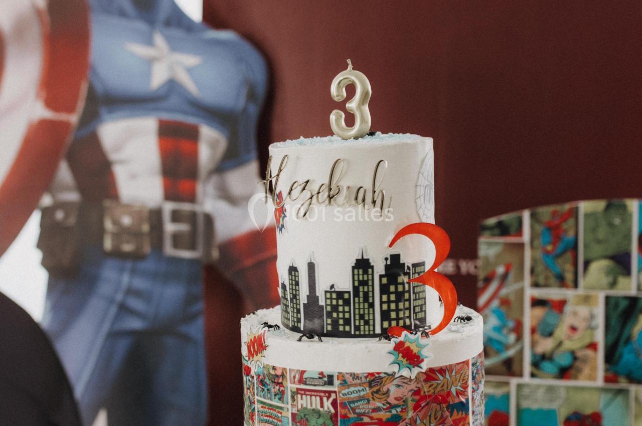 Gâteau d'anniversaire à trois étages décoré sur le thème des super-héros, avec un chiffre 3 au sommet.
