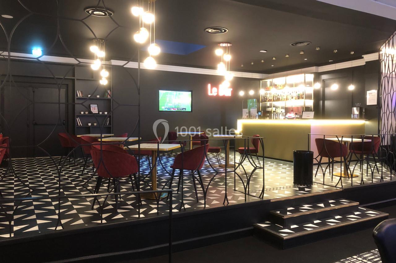 Espace lounge moderne avec tables, chaises rouges, bar éclairé et écran diffusant une émission sportive.