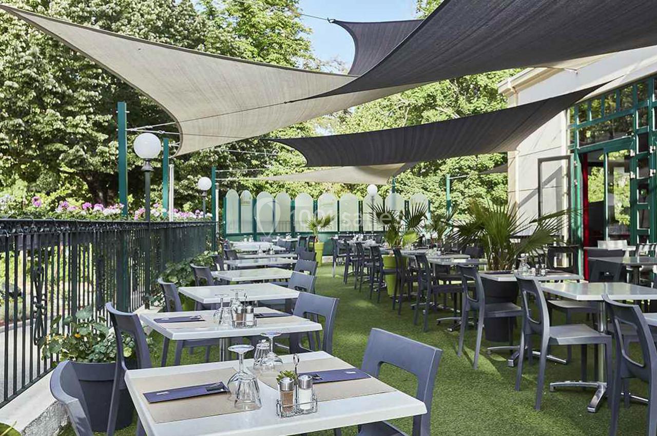 Terrasse de restaurant avec tables et chaises grises, ombragée par des voiles tendues, entourée de verdure.