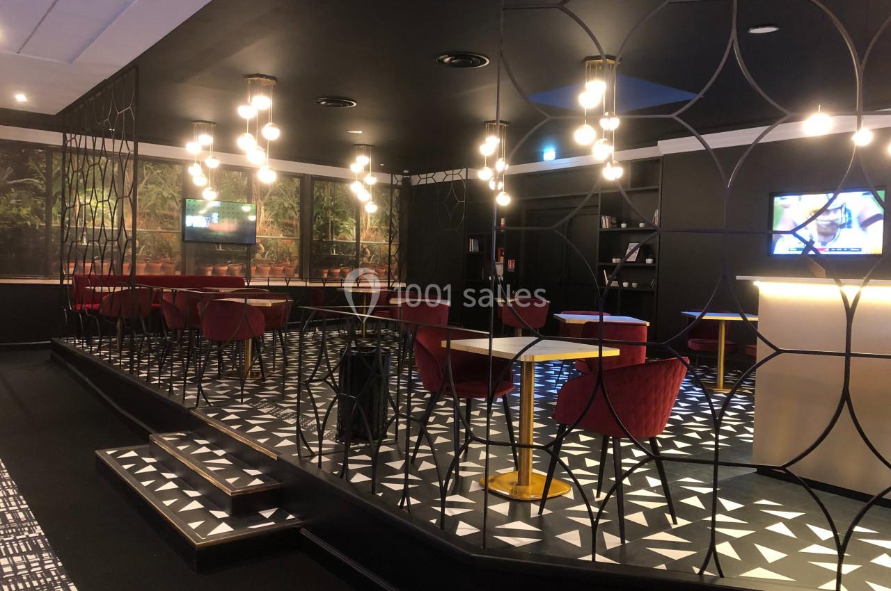 Espace lounge moderne avec tables, chaises rouges, éclairage suspendu et écran allumé sur un mur sombre.
