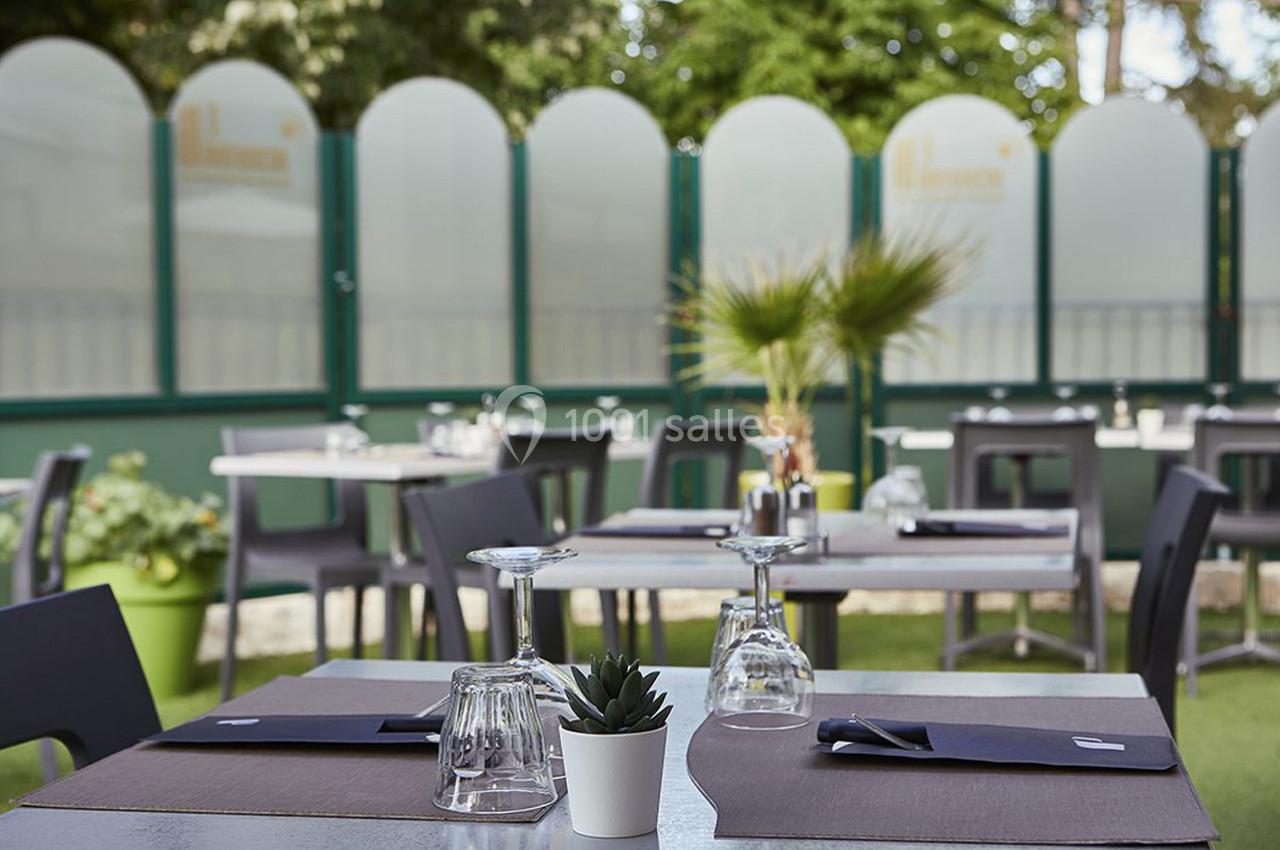 Tables dressées en extérieur avec nappes sombres, verres et plantes, entourées de panneaux translucides et verdure.