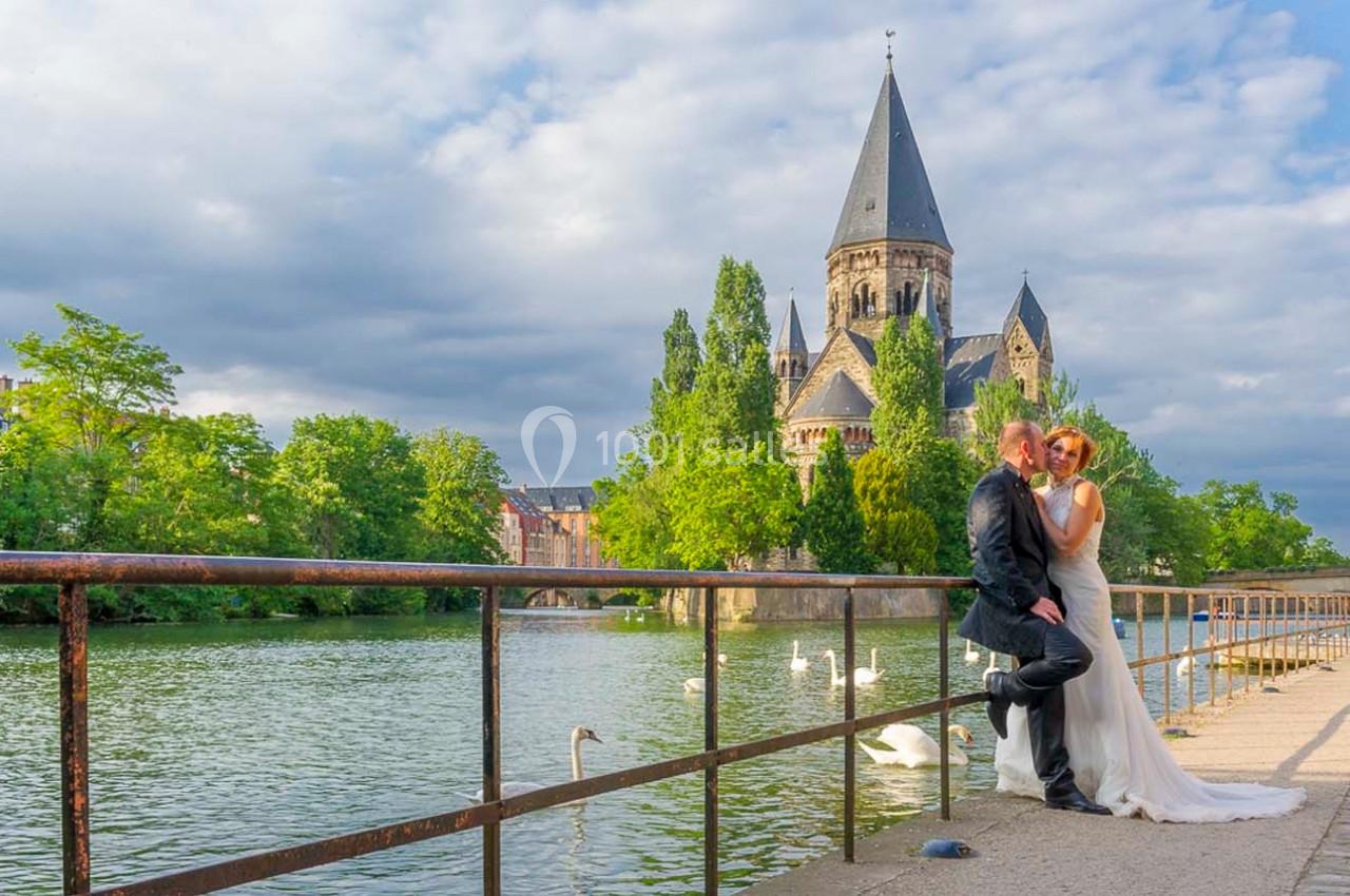 Un couple en tenue de mariage pose près d'une balustrade avec une église et une rivière en arrière-plan.