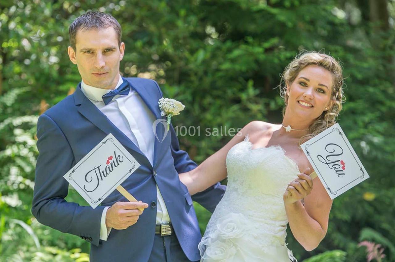 Un couple en tenue de mariage tient des pancartes ’Thank’ et ’You’ dans un cadre verdoyant.