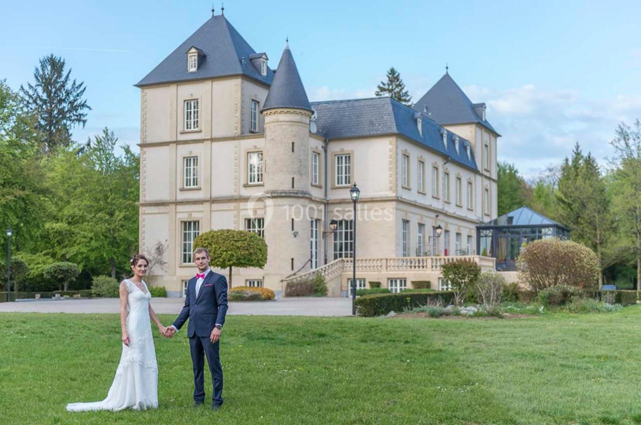 Un couple en tenue de mariage pose devant un château entouré de verdure par une journée ensoleillée.