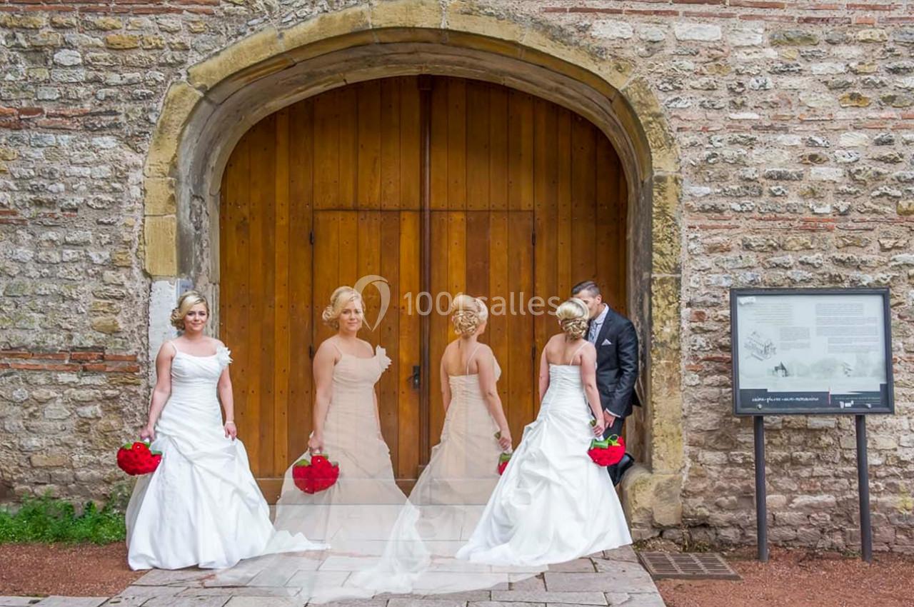 Quatre femmes en robe de mariée et un homme en costume posent devant une grande porte en bois et un mur en pierre.