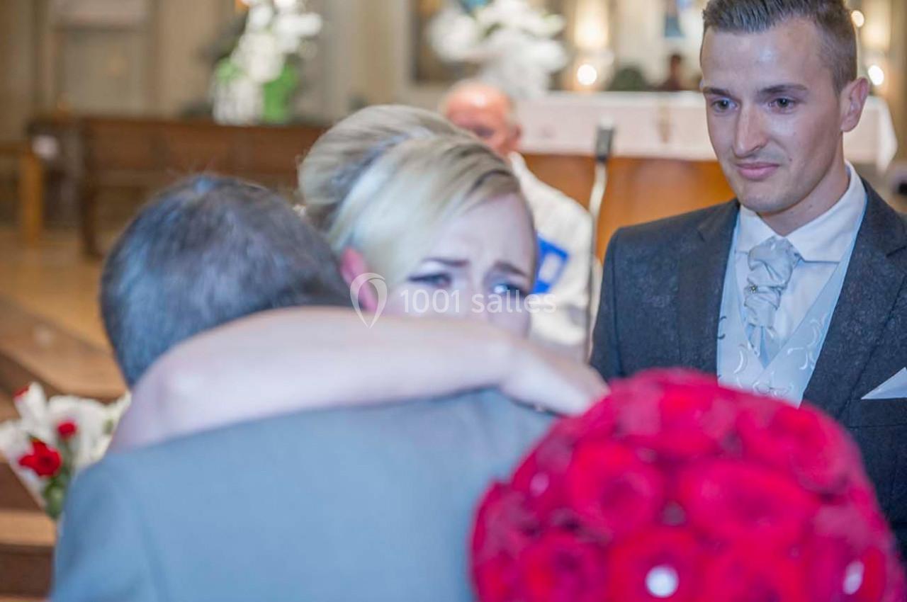 Une femme émue enlace un homme dans une église, tenant un bouquet de roses rouges, avec un autre homme en arrière-plan.