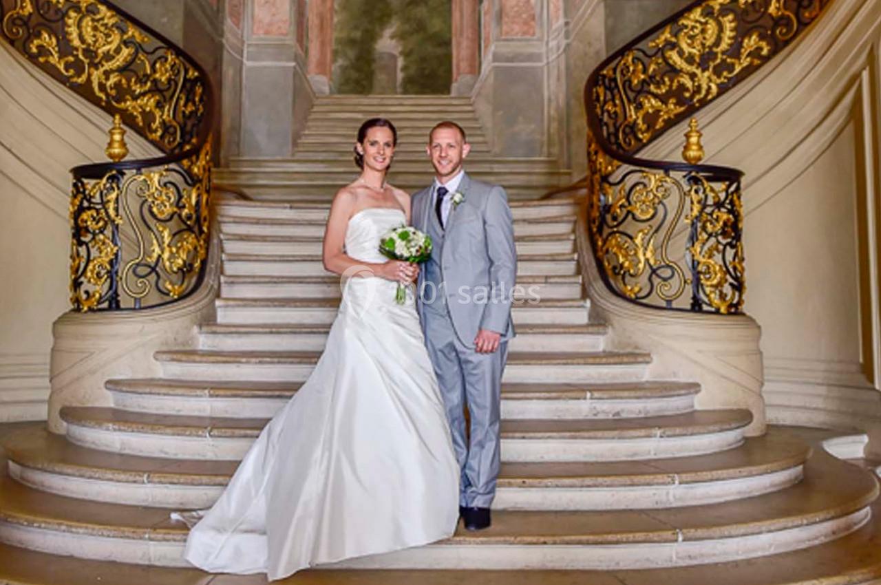 Un couple en tenue de mariage pose sur un escalier orné de rampes dorées dans un décor élégant.