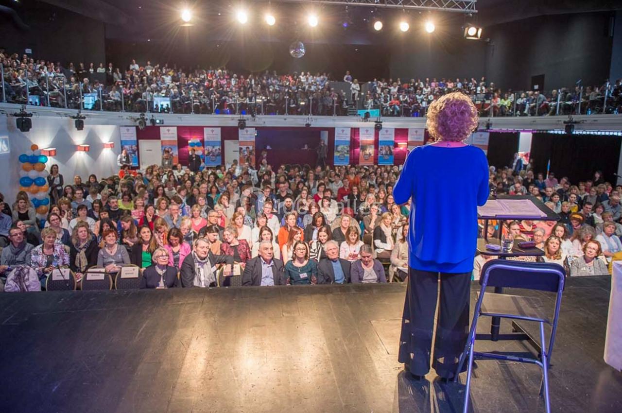 Une personne s'adresse à un large public assis dans une salle de conférence éclairée.