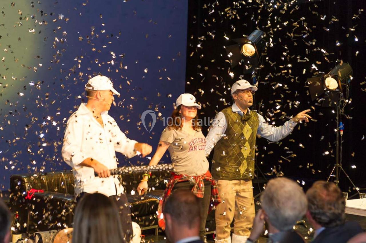 Trois personnes sur scène sous des projecteurs, entourées de confettis, devant un public.