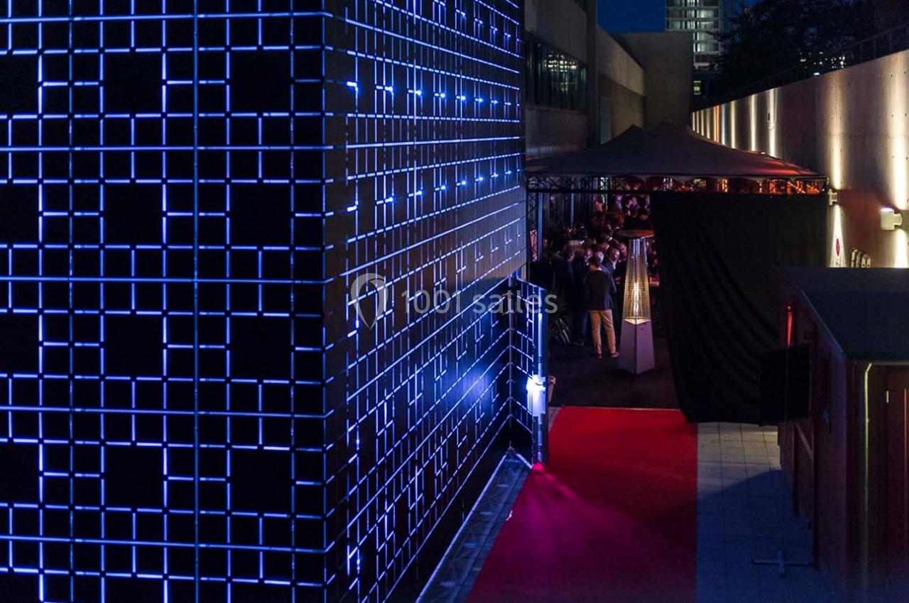 Façade illuminée par des motifs géométriques bleus, avec un tapis rouge menant à une entrée sous un auvent.