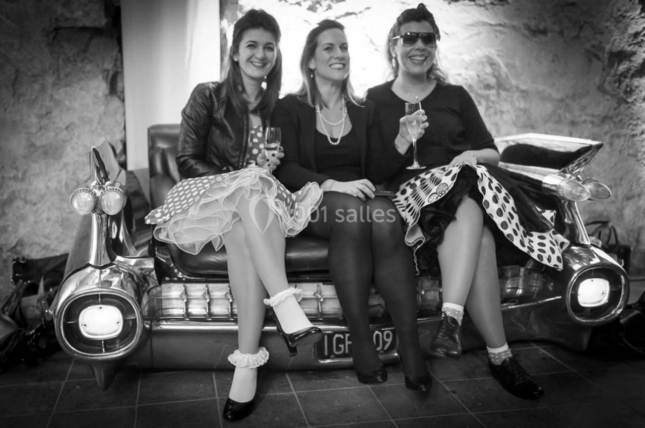 Trois femmes assises sur un canapé en forme de voiture rétro, tenant des verres, dans un décor intérieur en noir et blanc.