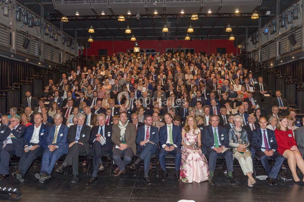 Un large groupe de personnes assises dans un auditorium, regardant vers l'objectif, sous un éclairage intérieur.