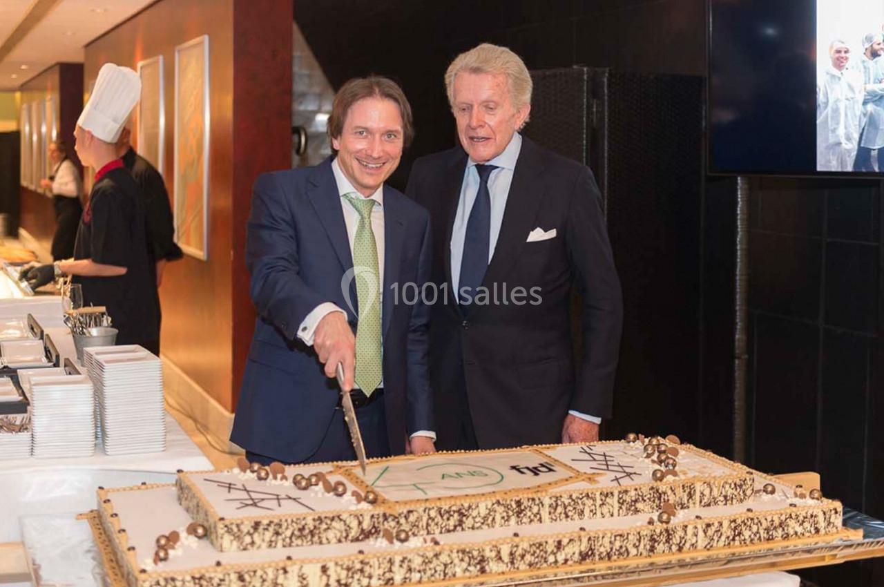 Deux hommes en costume coupent un grand gâteau décoré, dans un cadre formel avec un chef en arrière-plan.