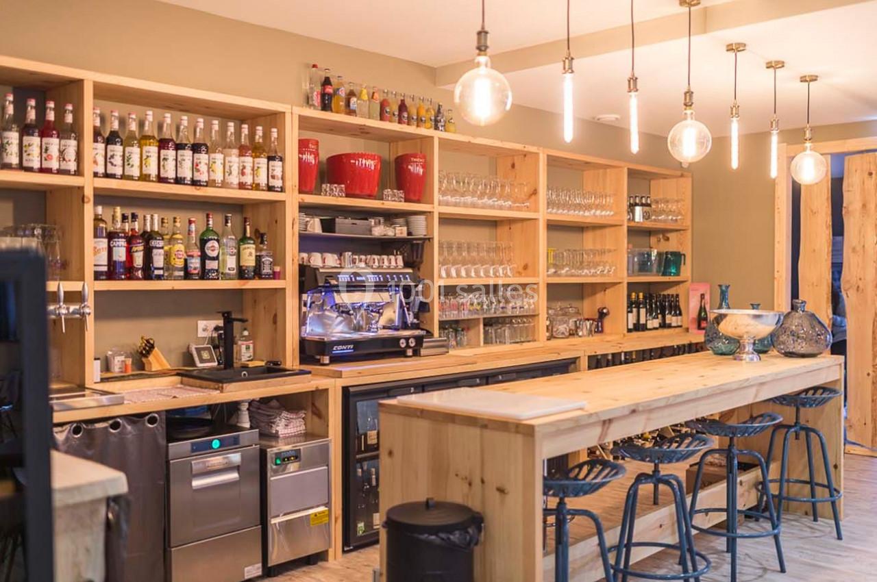 Bar en bois avec étagères remplies de bouteilles et verres, machine à café et tabourets alignés au comptoir.