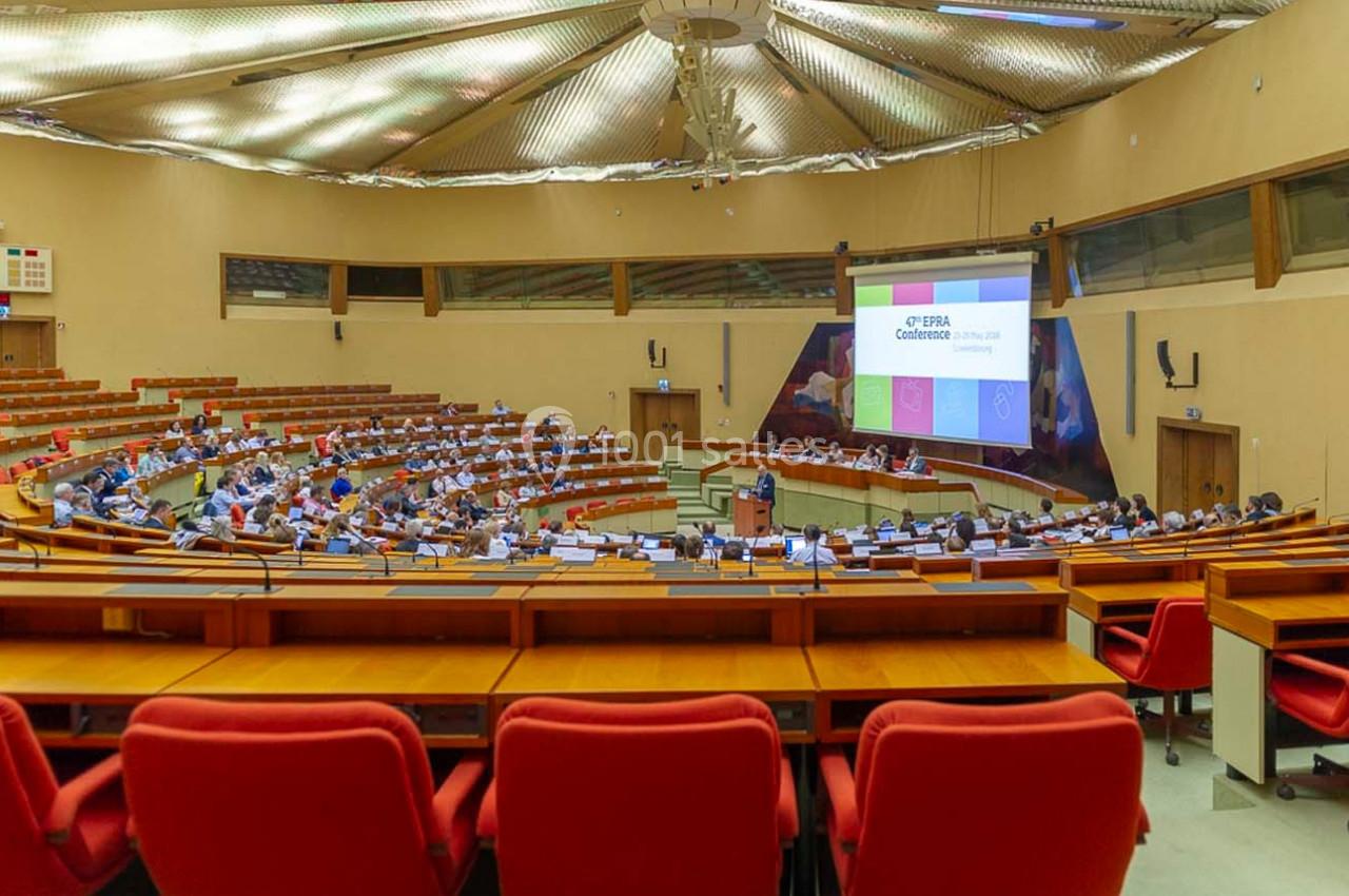 Salle de conférence avec des participants assis, un orateur debout et une présentation projetée à l'écran.