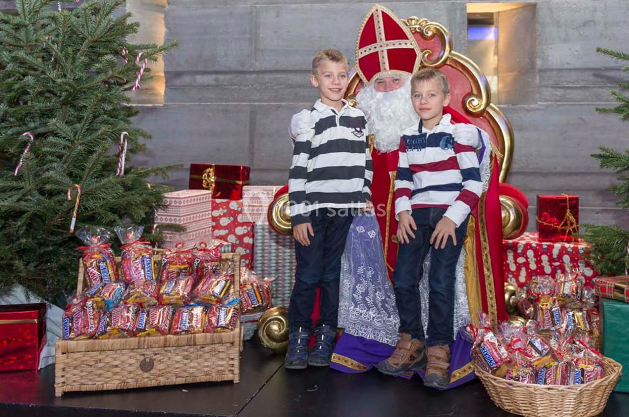 Deux enfants posent avec Saint-Nicolas sur un fauteuil rouge, entourés de sapins, cadeaux et paniers de friandises.