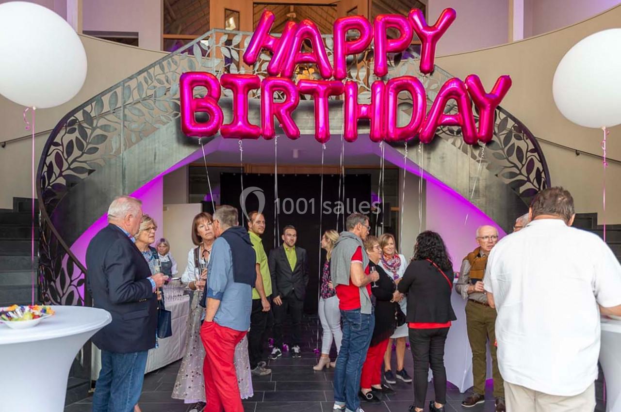 Groupe de personnes réunies dans un hall décoré avec des ballons roses formant ’Happy Birthday’.