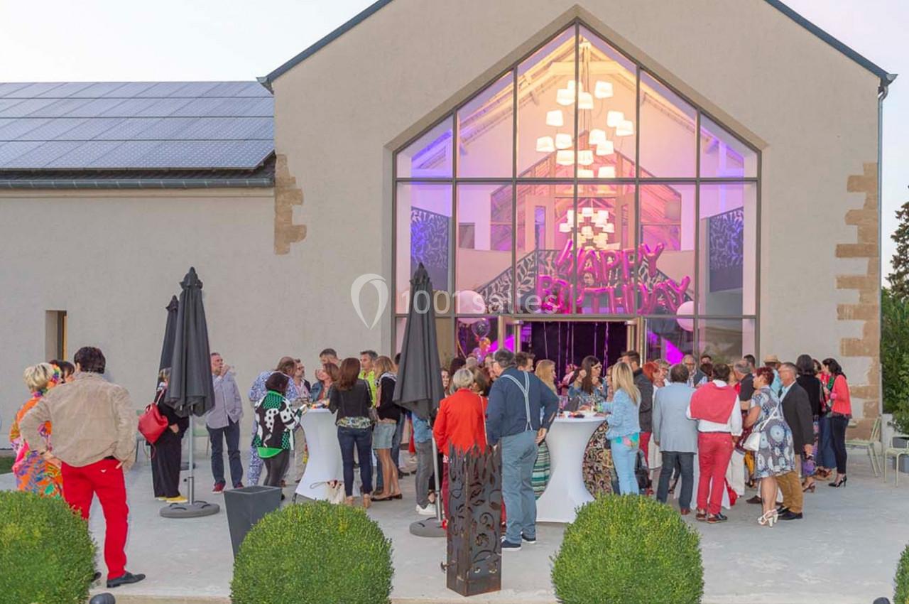 Groupe de personnes rassemblées devant une salle vitrée illuminée, décorée pour une fête d'anniversaire.