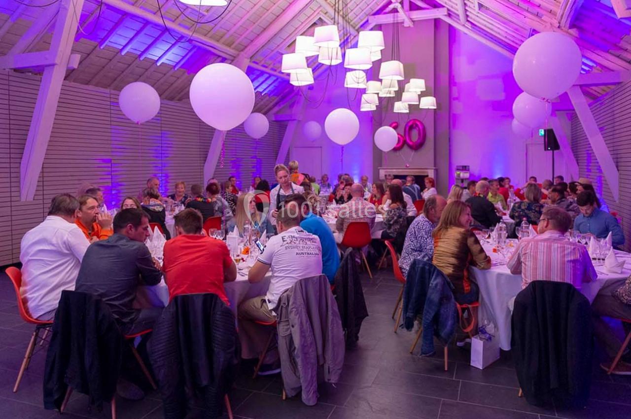 Salle décorée avec des lumières violettes, des ballons suspendus et des invités assis à des tables lors d'un événement.