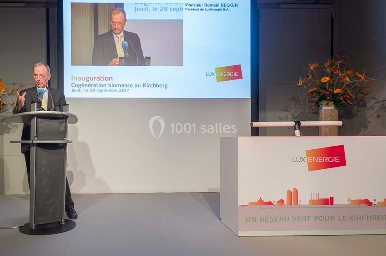 Un homme en costume parle à un pupitre lors d'une inauguration, avec un écran et un stand Lux Énergie en arrière-plan.