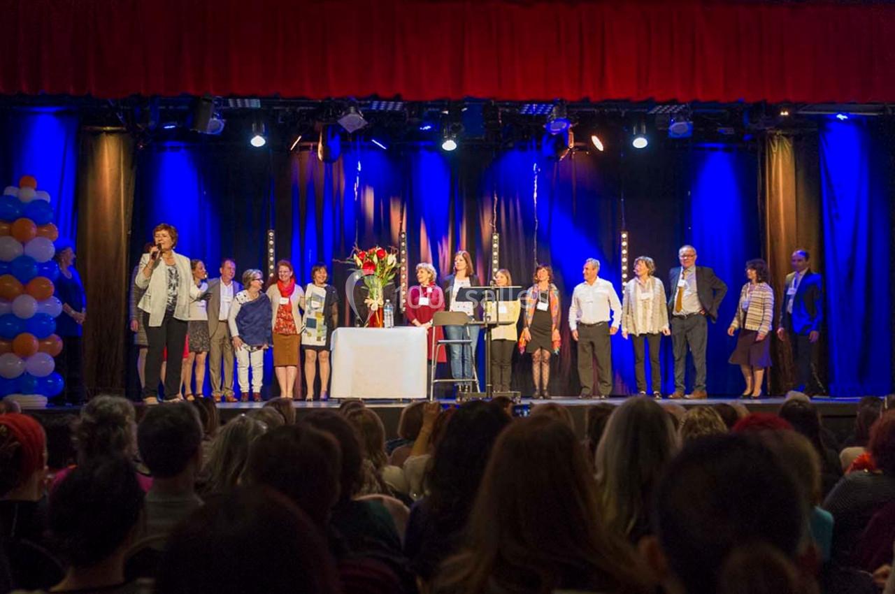 Un groupe de personnes debout sur une scène éclairée, face à un public, avec des rideaux rouges et bleus en arrière-plan.
