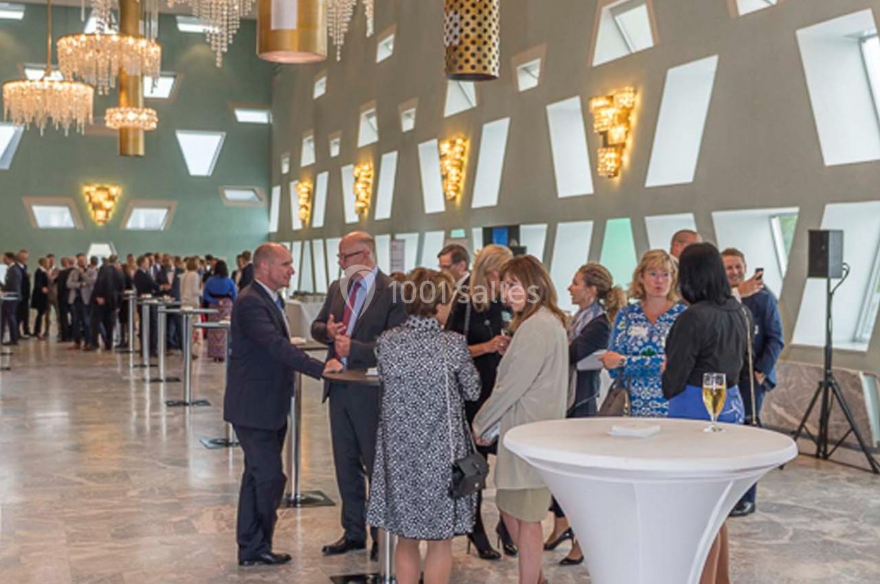 Des personnes discutent debout dans une salle élégante avec des lustres imposants et des murs aux fenêtres géométriques.
