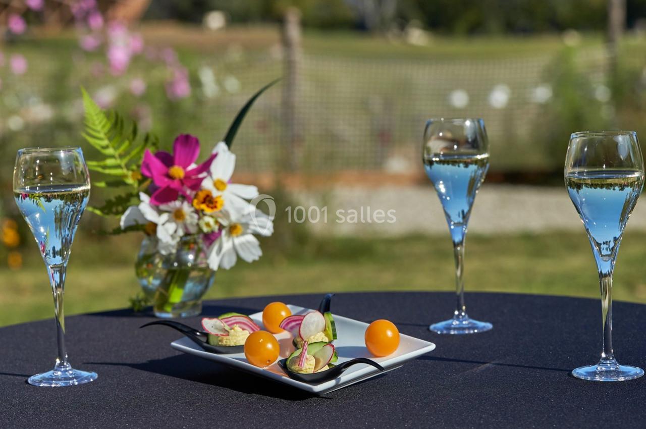 Trois verres de vin blanc et une assiette de bouchées colorées sur une table noire en extérieur, avec fleurs en arrière-plan.