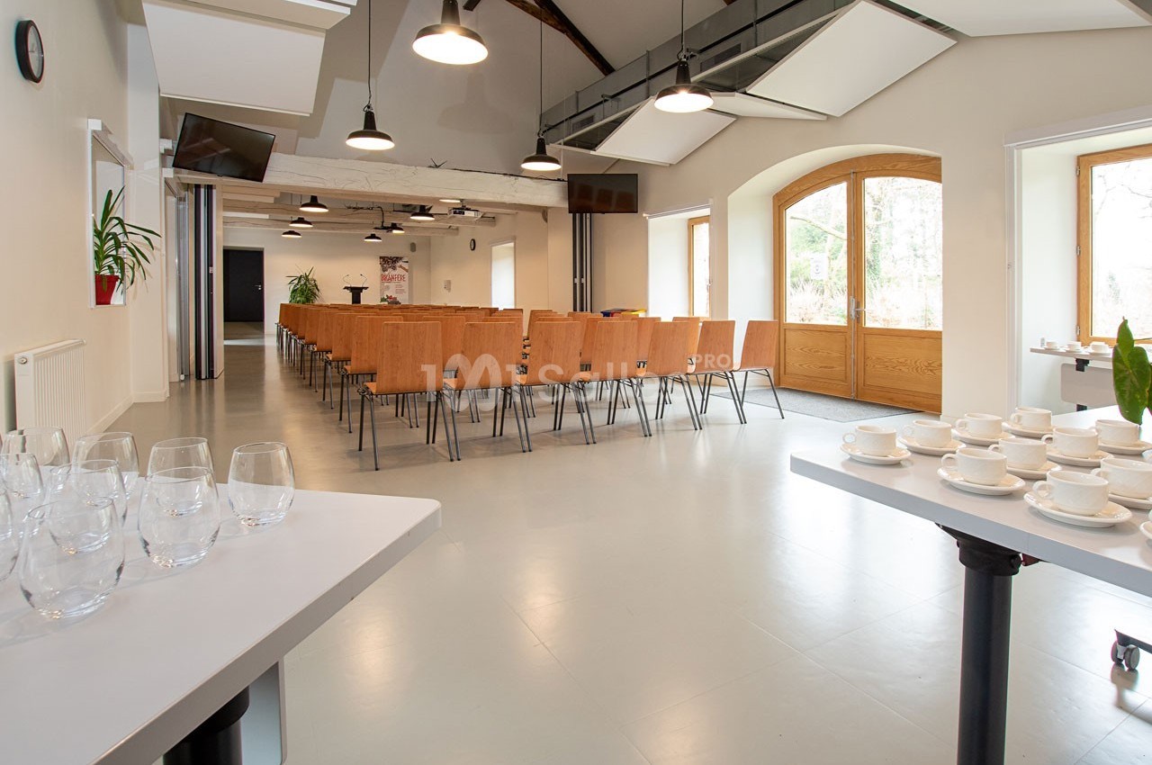 Salle lumineuse avec chaises alignées, tables dressées avec verres et tasses, grandes fenêtres et poutres apparentes.