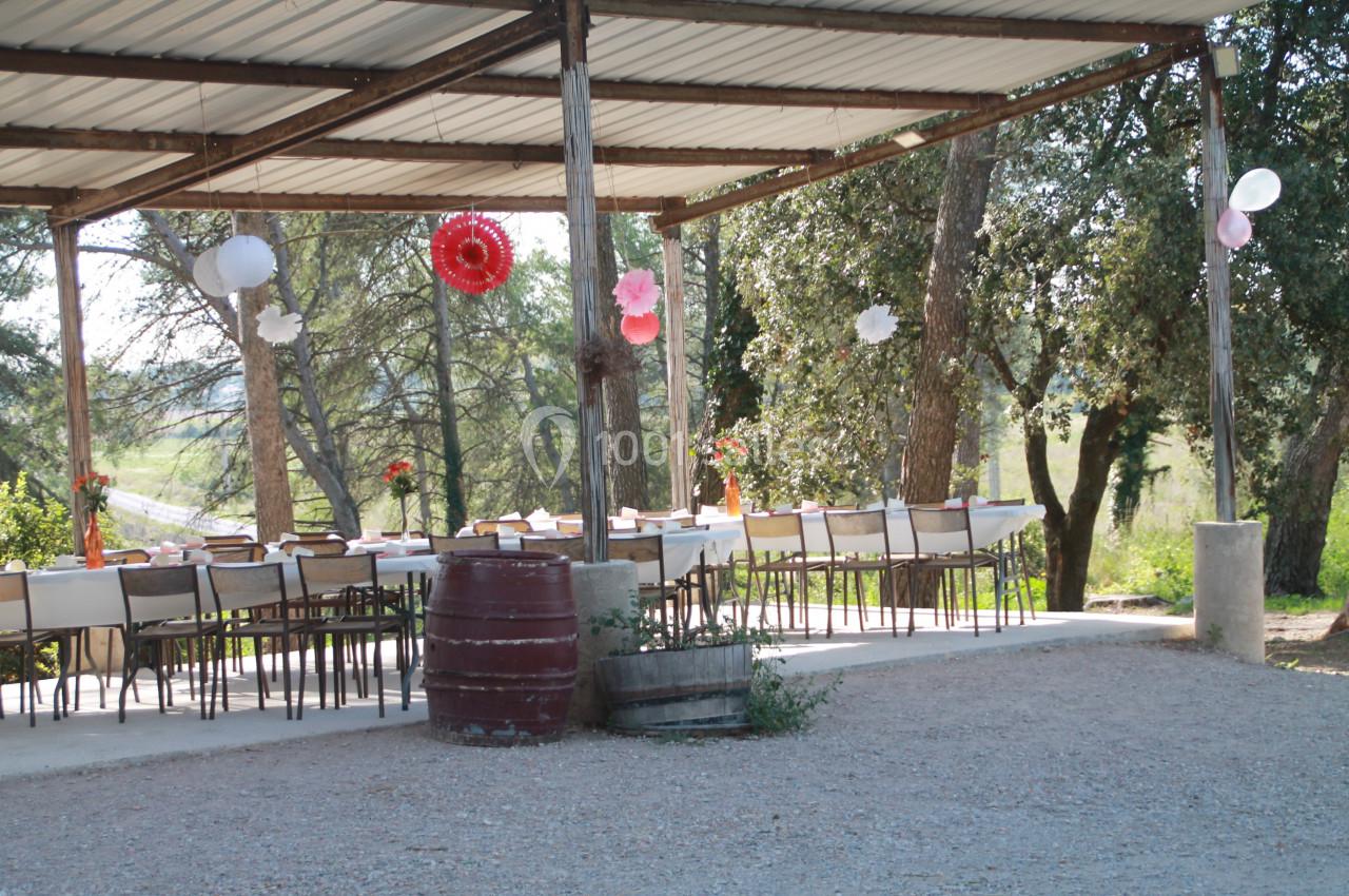 Table dressée sous une pergola en plein air, décorée de lanternes et fleurs, entourée d'arbres et de végétation.