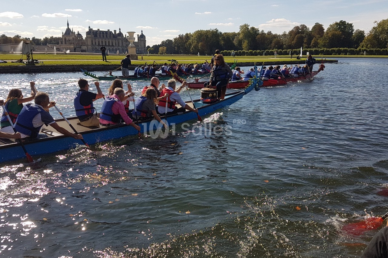 Des équipes pagaient sur des bateaux-dragons dans un plan d'eau entouré de verdure et d'un château en arrière-plan.