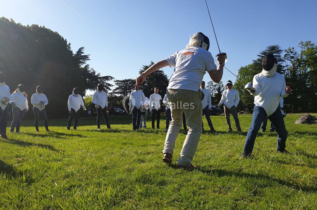 Un groupe de personnes en tenue d'escrime s'entraîne en plein air sur une pelouse par une journée ensoleillée.