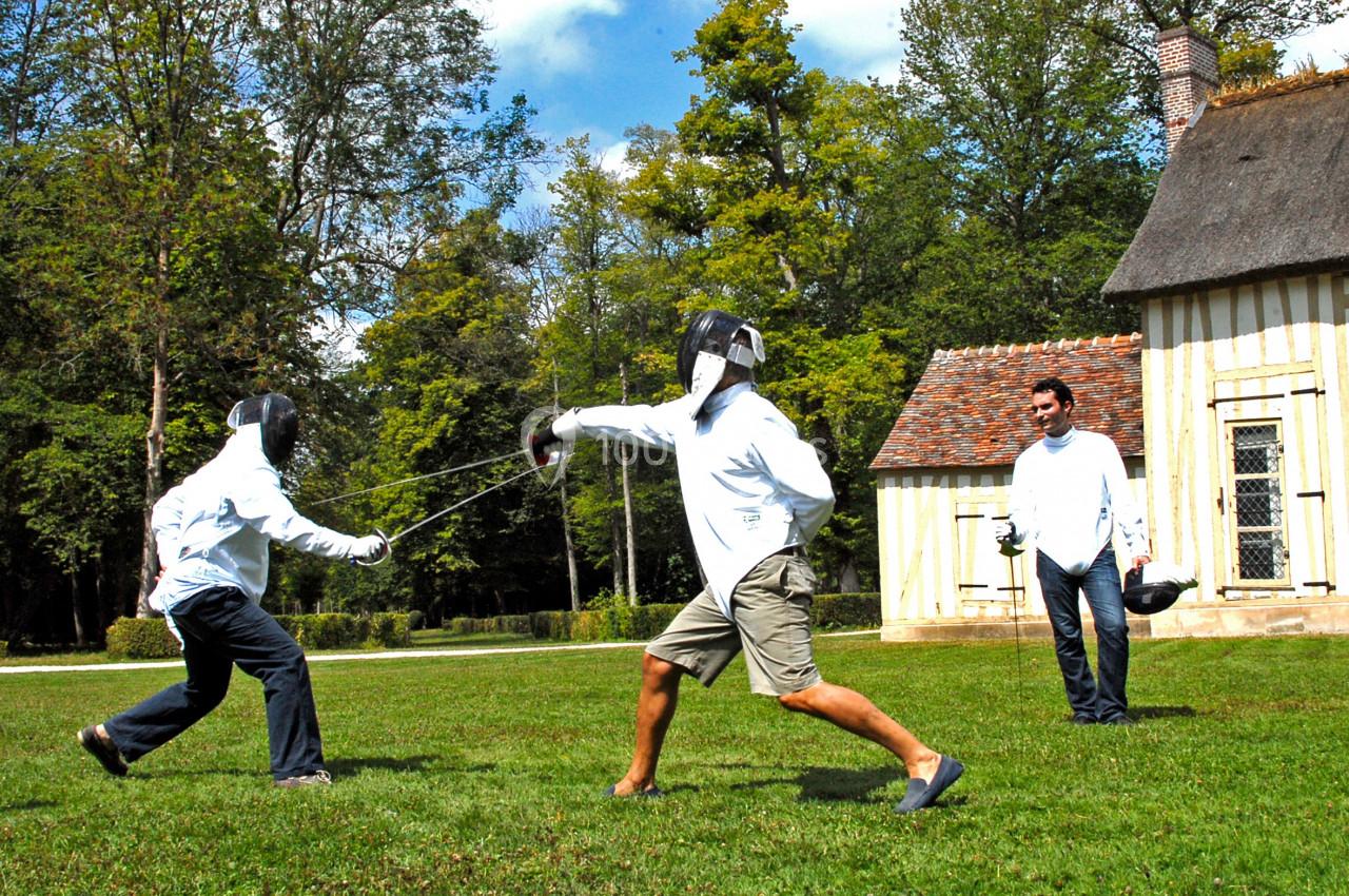 Deux personnes pratiquent l'escrime en plein air sur une pelouse, sous le regard d'un spectateur près d'une maison.