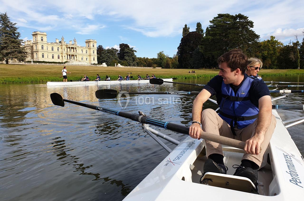 Des personnes rament sur un lac calme près d'un grand bâtiment historique entouré de verdure.
