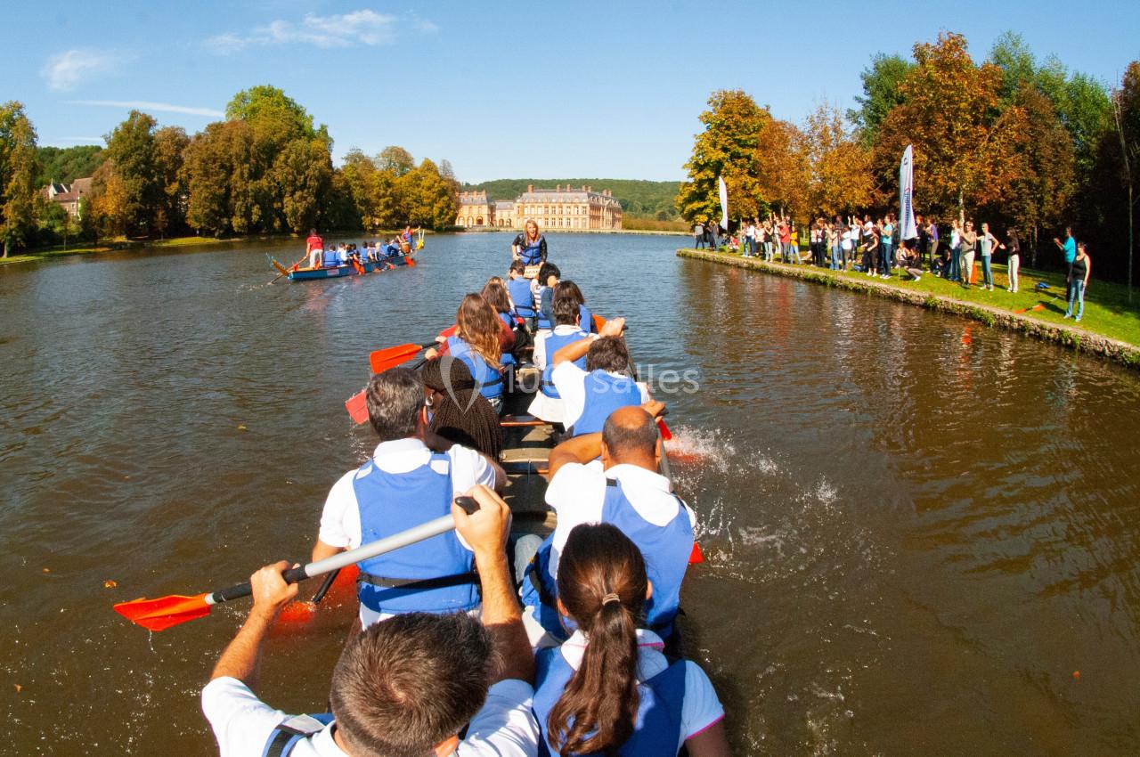 Des rameurs en gilets bleus pagaient sur un lac bordé d'arbres, sous un ciel dégagé, avec des spectateurs sur la rive.
