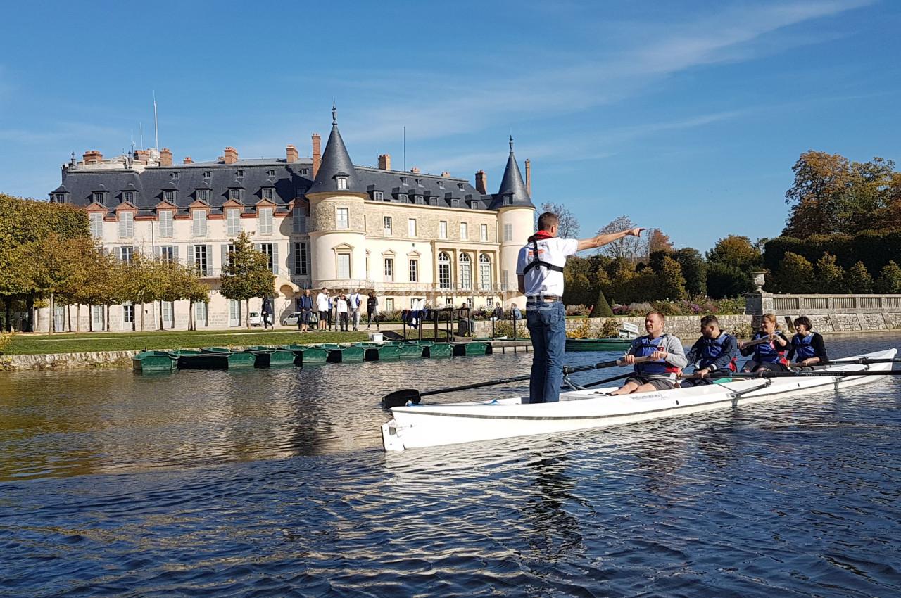 Un groupe pratique l'aviron sur une rivière devant un château entouré d'arbres en automne.