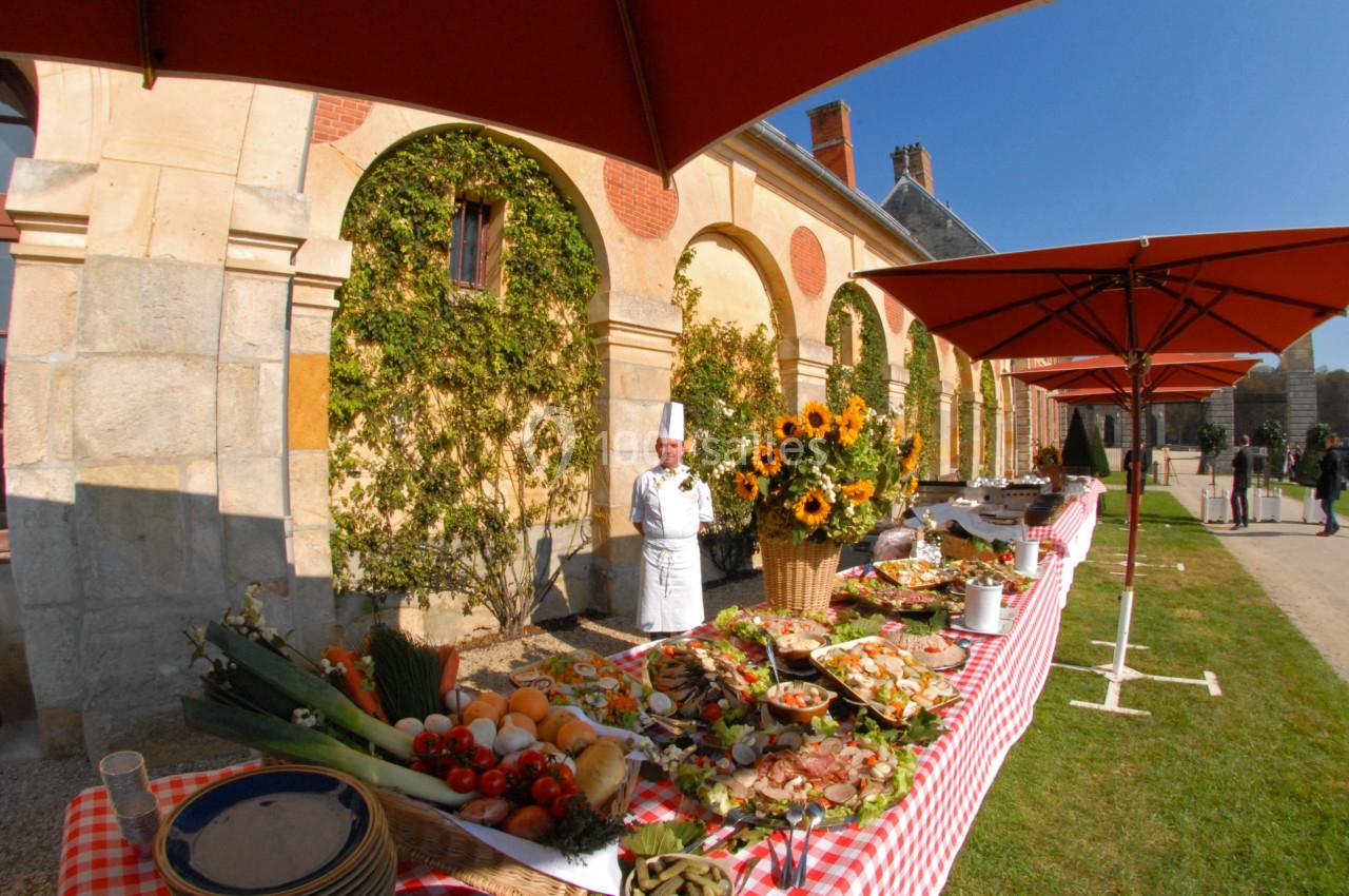 Buffet en plein air avec un chef devant une table garnie de plats variés, près d'un bâtiment en pierre et sous des parasols…