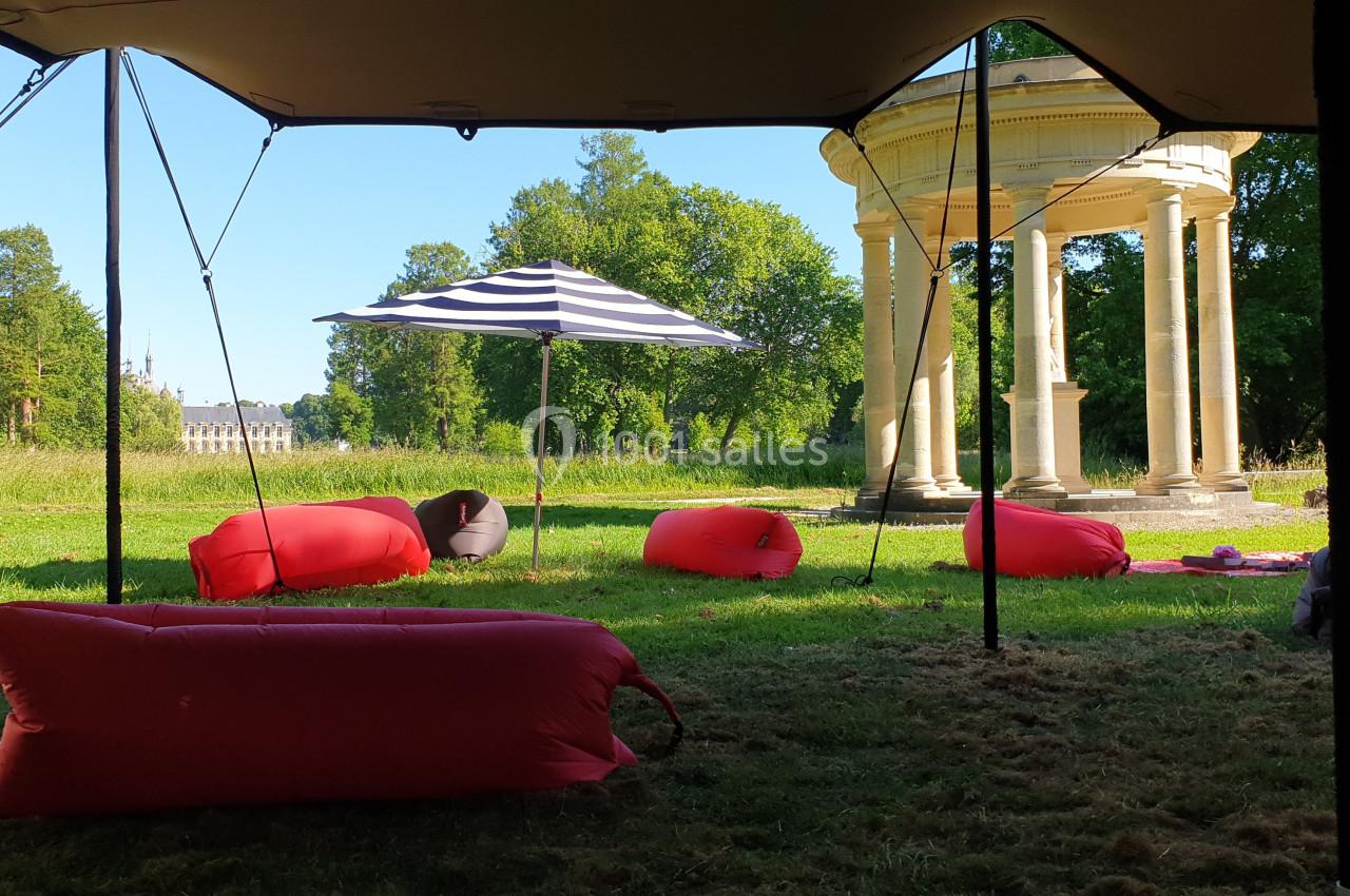 Des poufs colorés et un parasol rayé sont disposés sur une pelouse près d'un kiosque dans un parc verdoyant.