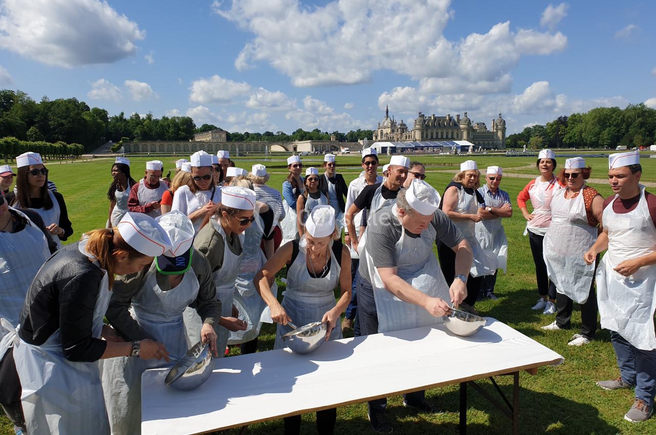 Un groupe de personnes en tabliers et toques cuisine en plein air sur une table, dans un cadre verdoyant et ensoleillé.
