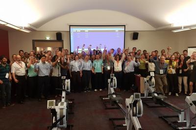 Un groupe de personnes souriantes pose dans une salle avec des rameurs et un écran projeté en arrière-plan.