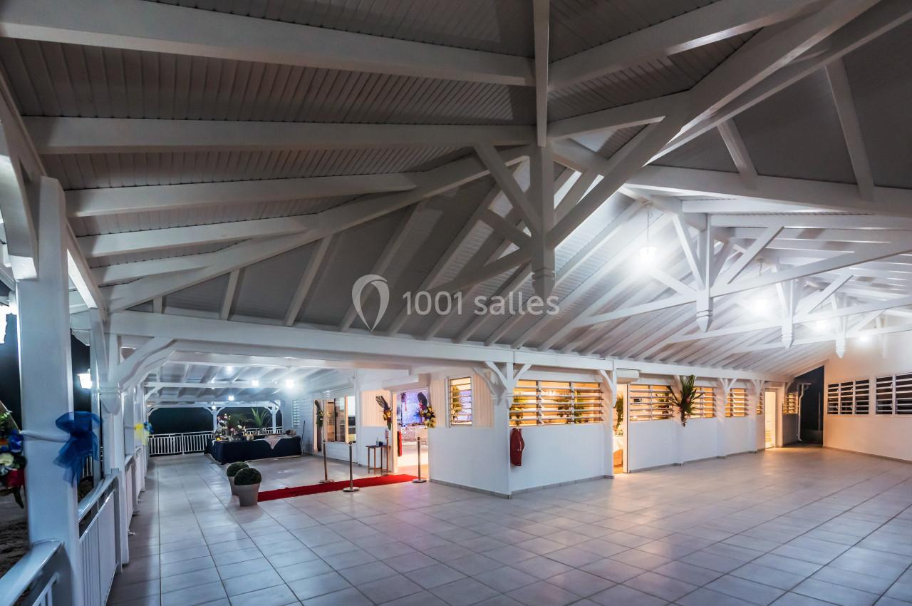 Grande salle couverte avec charpente en bois blanc, éclairage artificiel et sol carrelé clair.