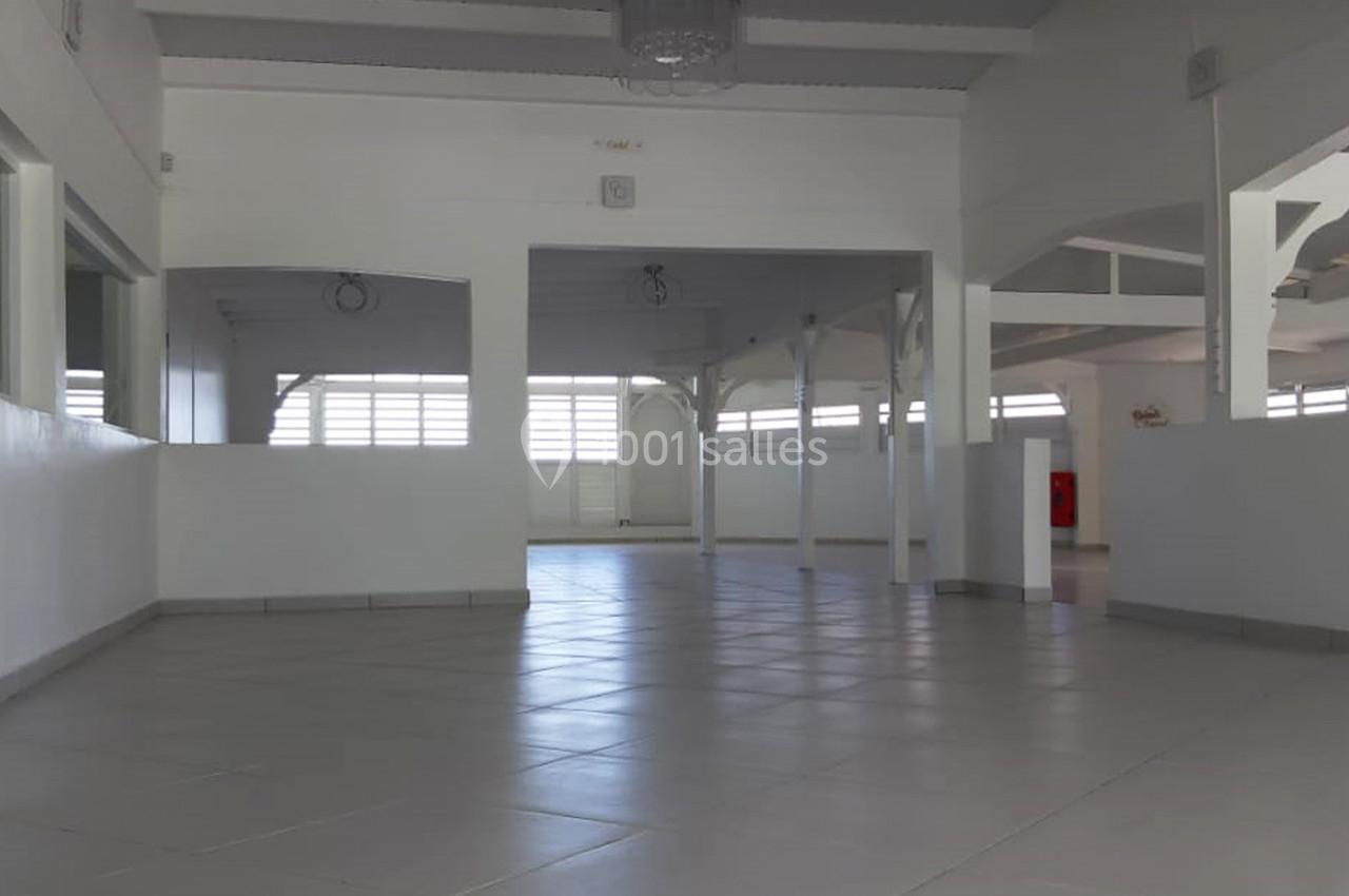 Salle vide avec sol carrelé, murs blancs, poutres apparentes et fenêtres laissant entrer la lumière naturelle.