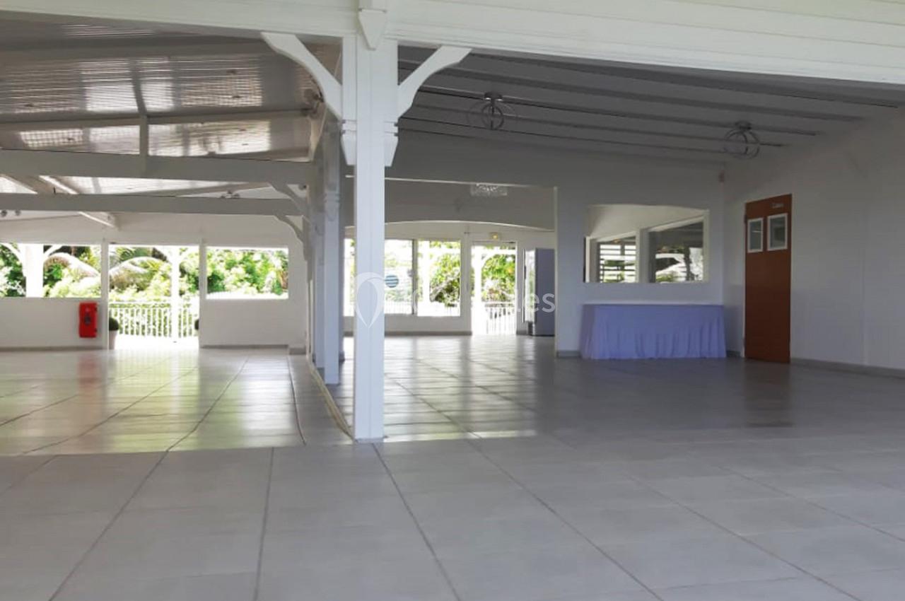 Salle lumineuse avec carrelage clair, grandes ouvertures vitrées, murs blancs et une table recouverte d'une nappe bleue.